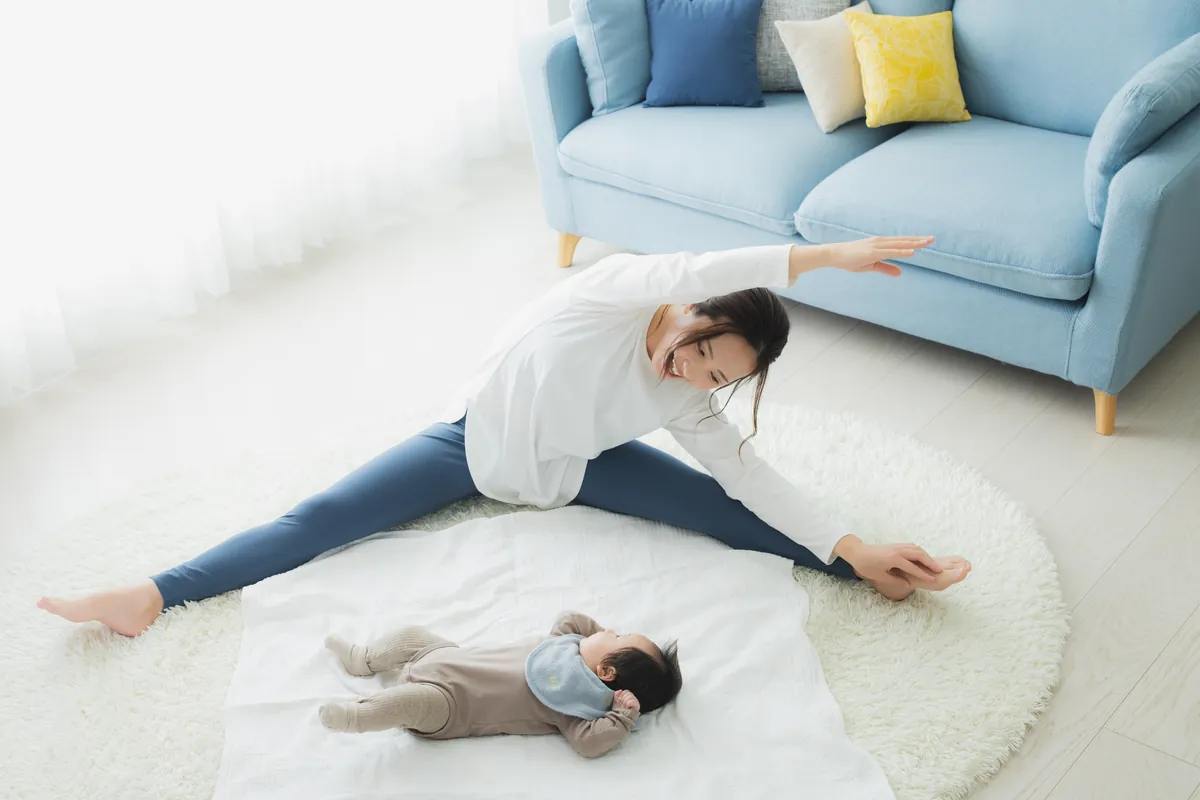自宅で赤ちゃんのそばでストレッチをする女性(イメージ)